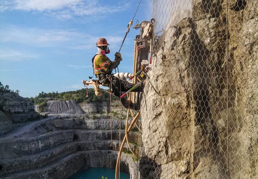 Worker on ropes installing mesh on a rock face above a quarry.