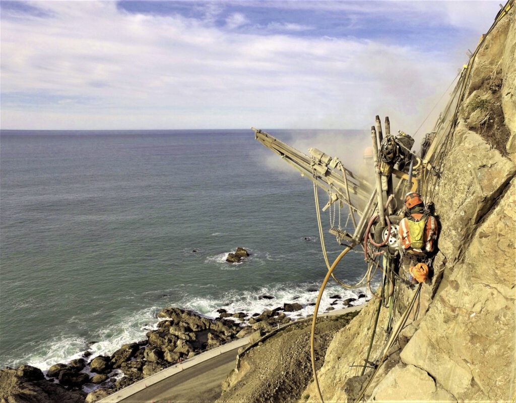 Worker on ropes operating a drill on a steep coastal cliff above a roadway.