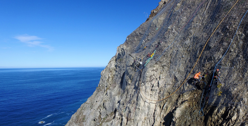 Rope access crew working on a steep coastal cliff face with ocean in the background.
