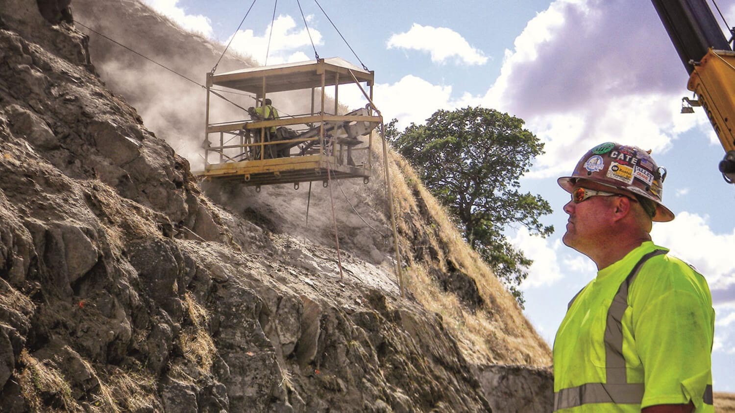 Construction worker observes as a suspended platform carries crew members up a steep rock slope.
