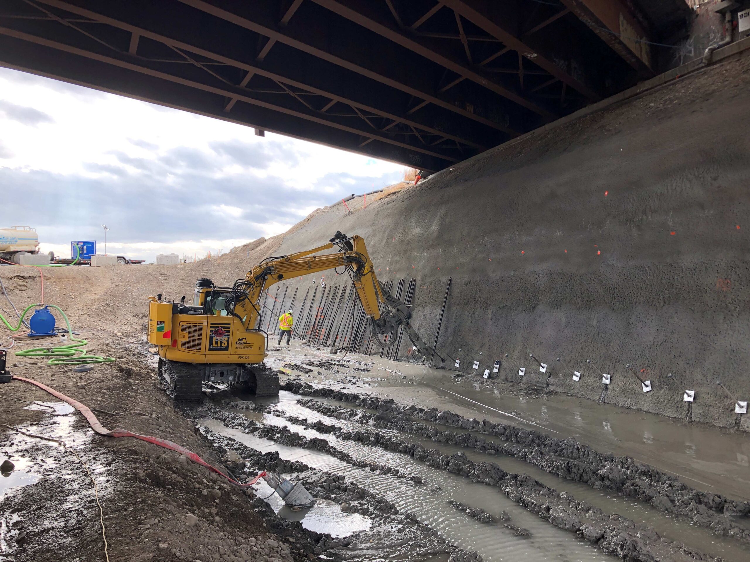 Excavator drilling into a shotcrete-covered slope beneath an overpass.